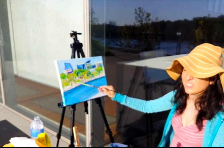 Woman painting on an easel outside, wearing a large sun hat, with a glass window reflecting trees and sky.