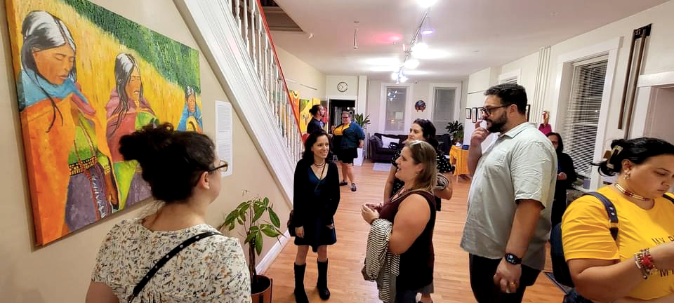People gather in an art gallery, viewing colorful paintings on the walls and engaging in conversation.