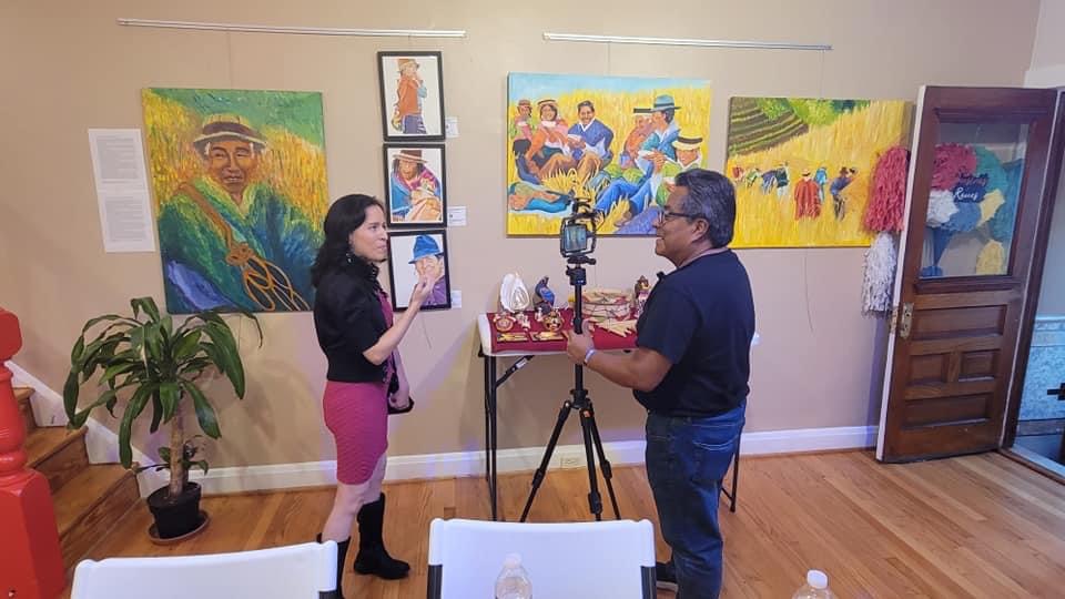 Two people talking in an art gallery with colorful paintings on the wall, a plant, and a camera on a tripod.