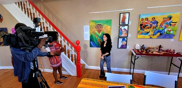 Person being filmed by a camera crew in an art gallery with colorful paintings on the wall.