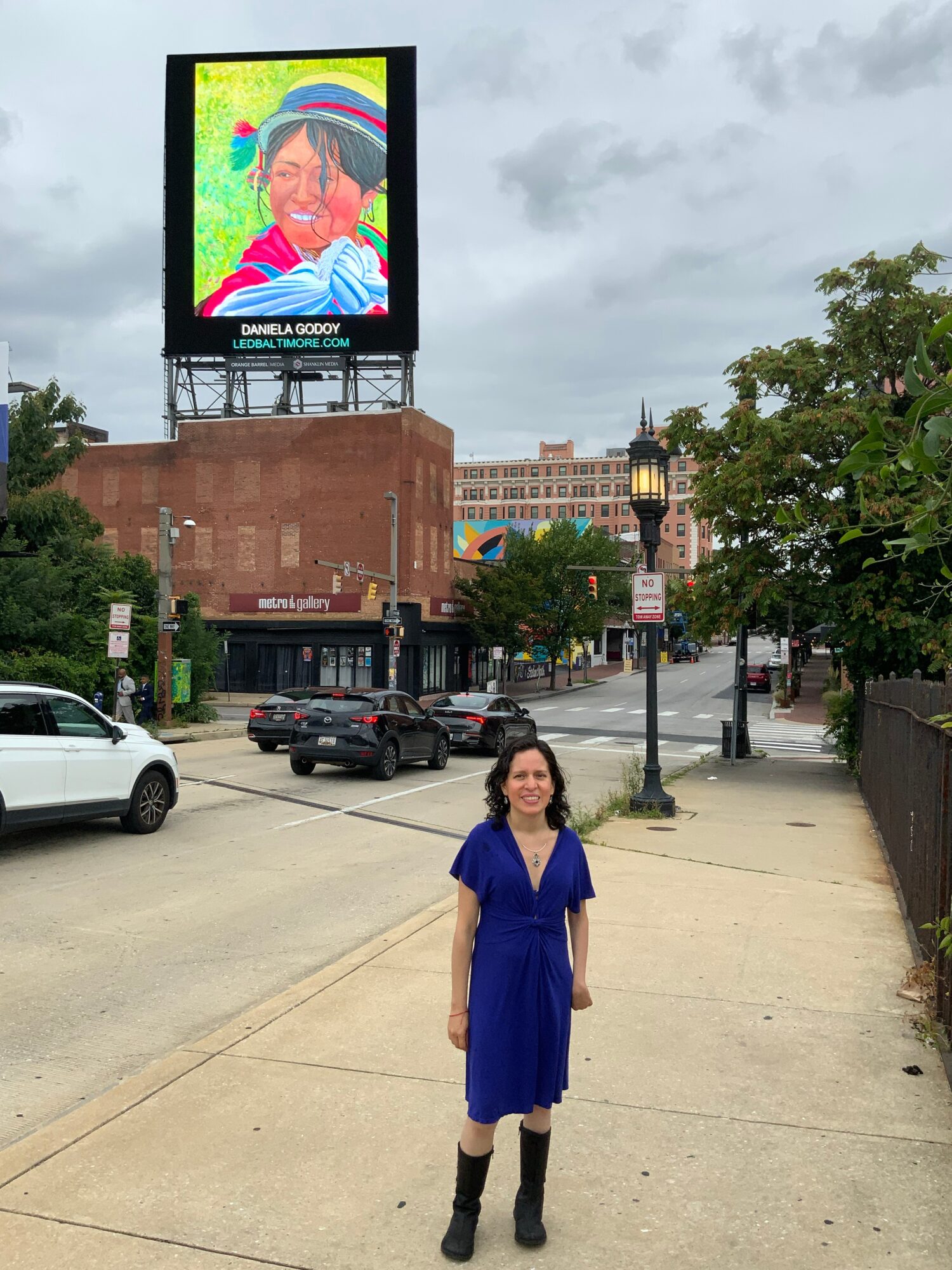 Woman in blue dress standing on sidewalk with city street, cars, and billboard in background.