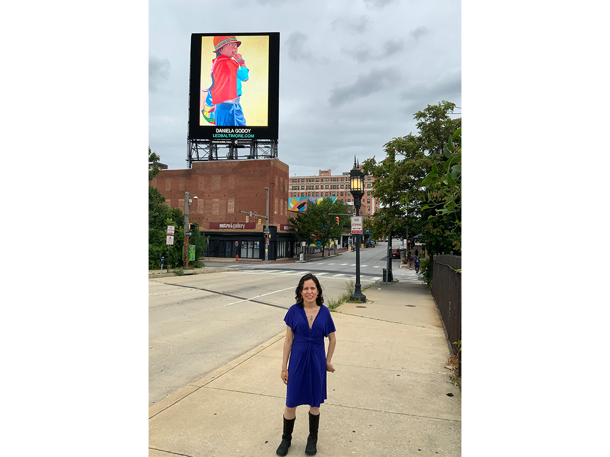 Woman in a blue dress standing on a city sidewalk with a large billboard and trees in the background.