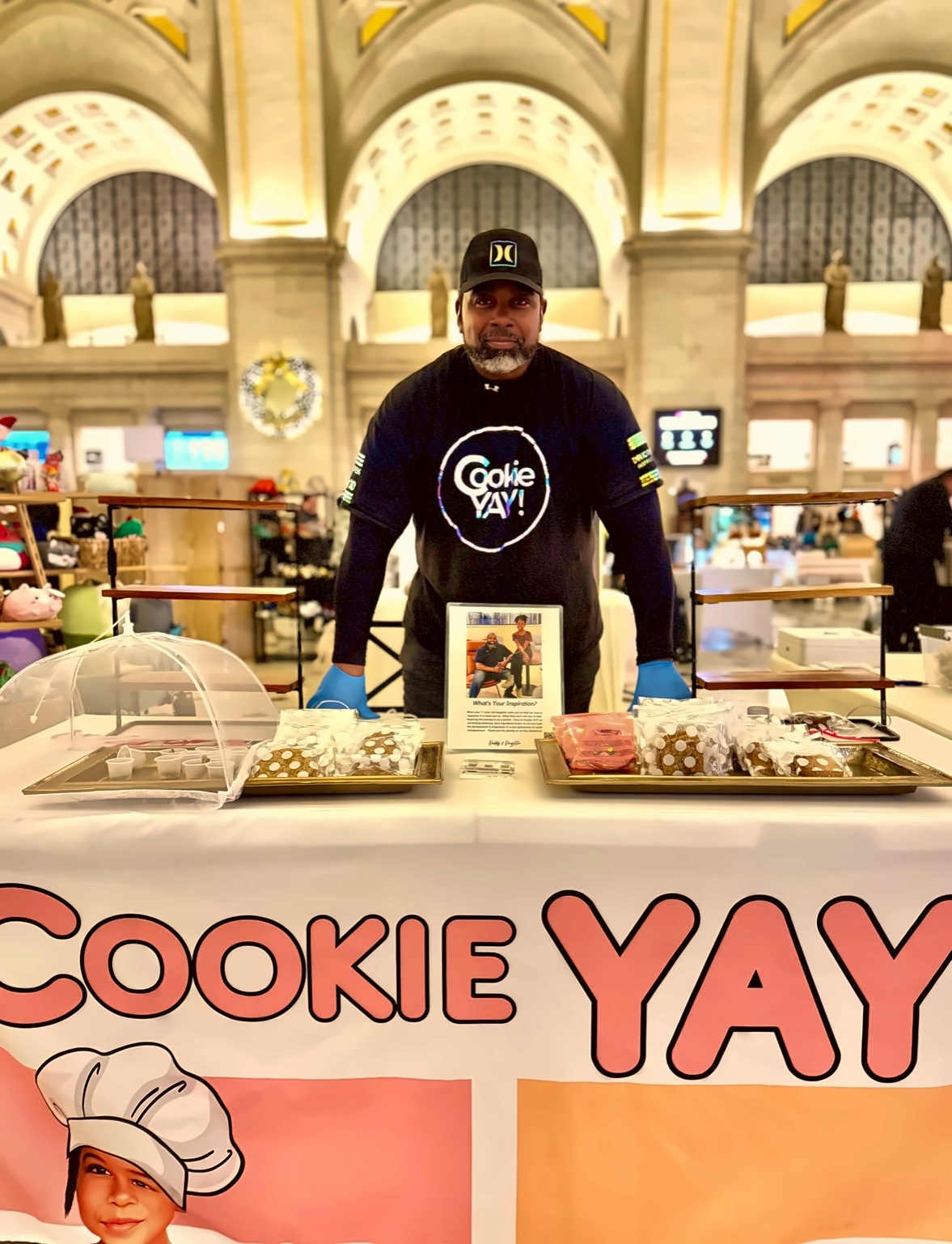 Man standing behind a cookie display counter in a large, ornate indoor space with arches and high ceilings.
