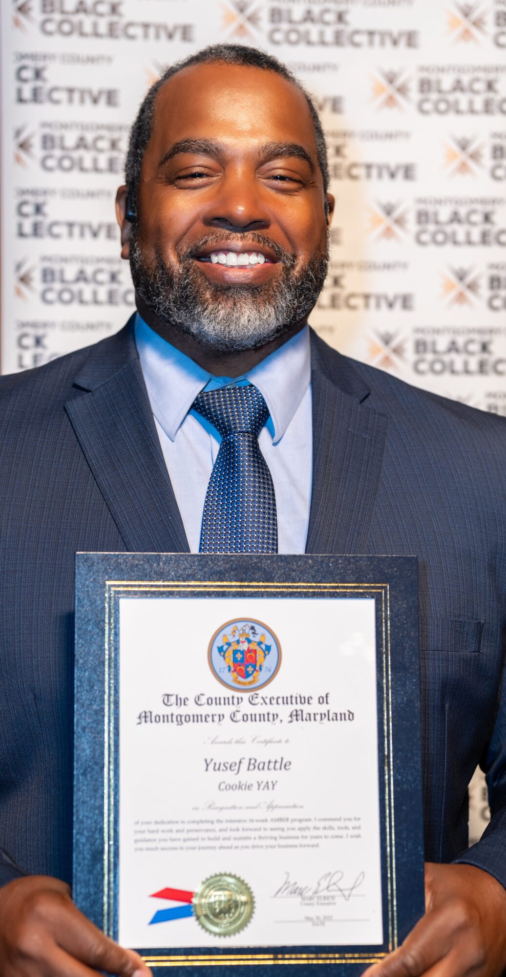 Man in suit holding a framed certificate, smiling, background with 'Black Collective' and 'Black Excellence' text