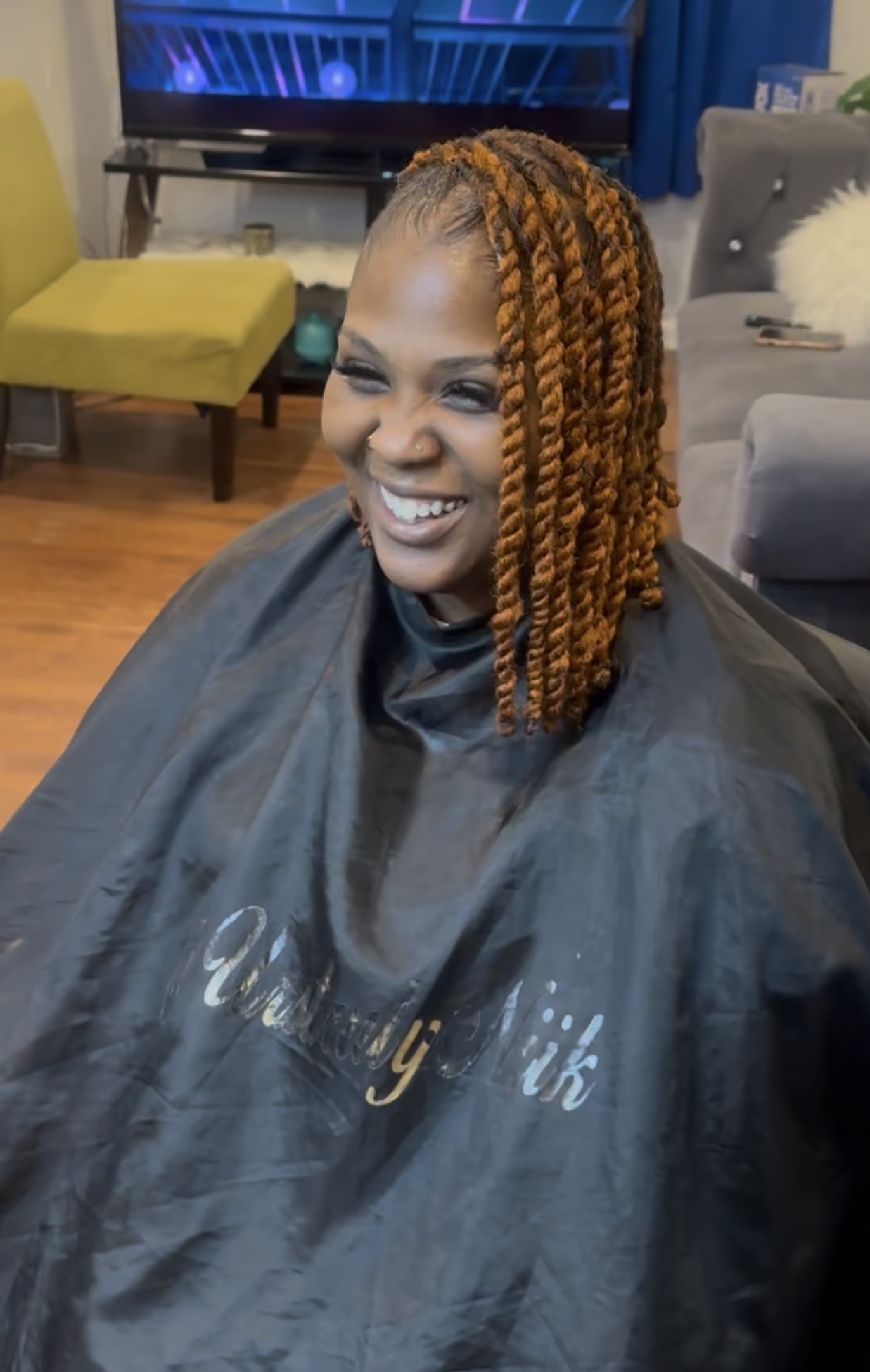 Woman with orange braids smiling in a salon chair, wearing a black cape, in a room with colorful furniture and a TV