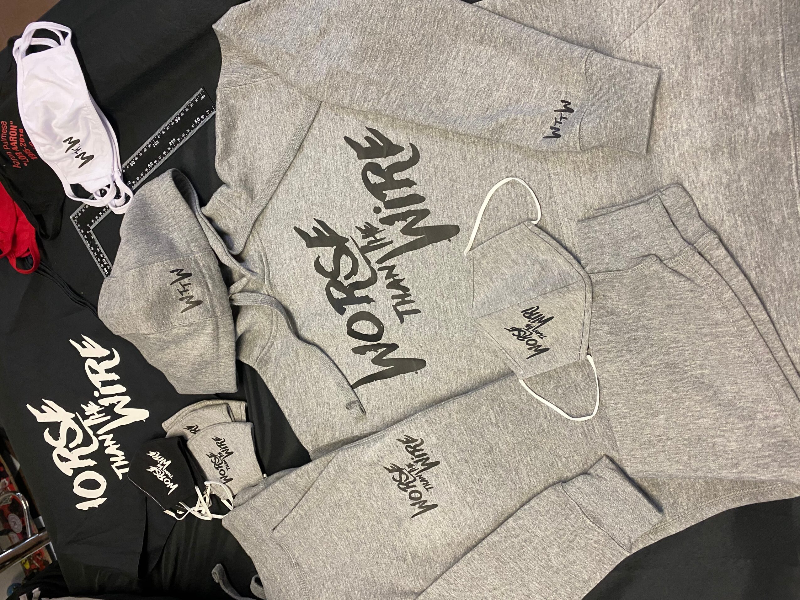 Gray hoodie with black text, gray sweatpants, and face mask on a gray surface, with clothing items nearby.