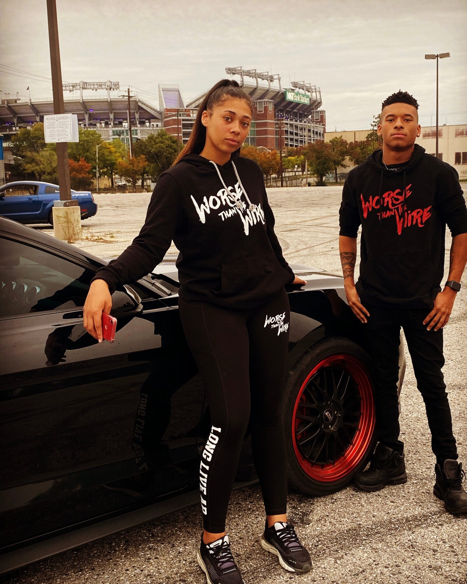 Two young people standing next to a black sports car with red wheels in a parking lot, cityscape background, woman leaning on car, man standing nearby