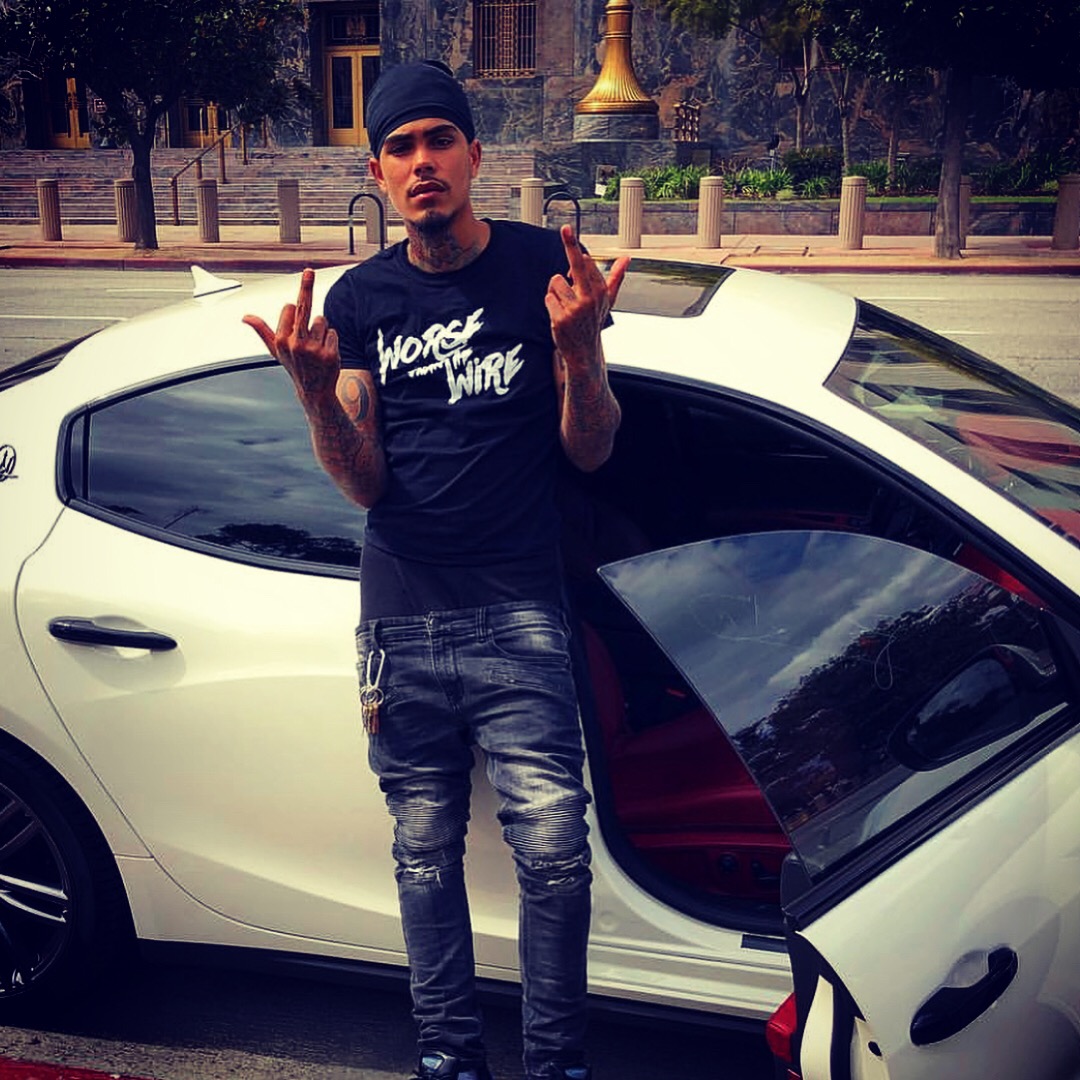 Young man standing next to a white car, making hand gestures, wearing a black beanie and casual clothing.