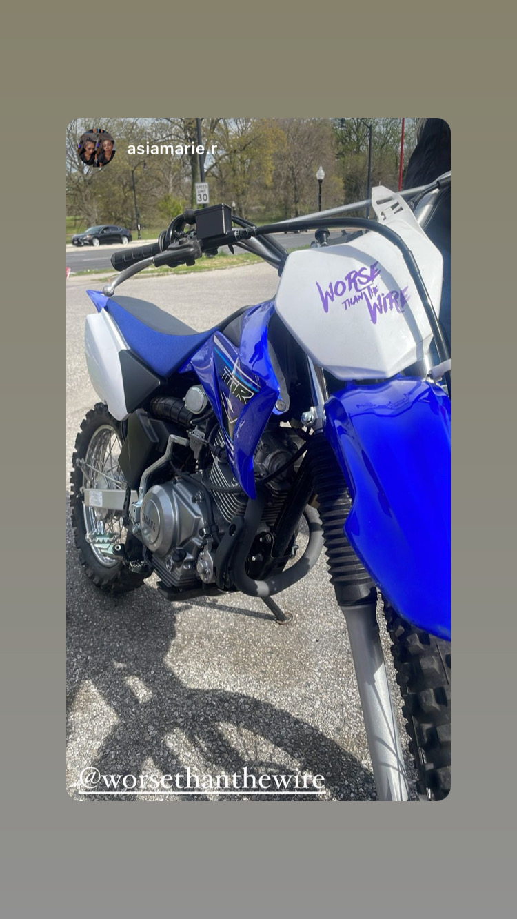Blue and white dirt bike parked outdoors with trees and a road in background.