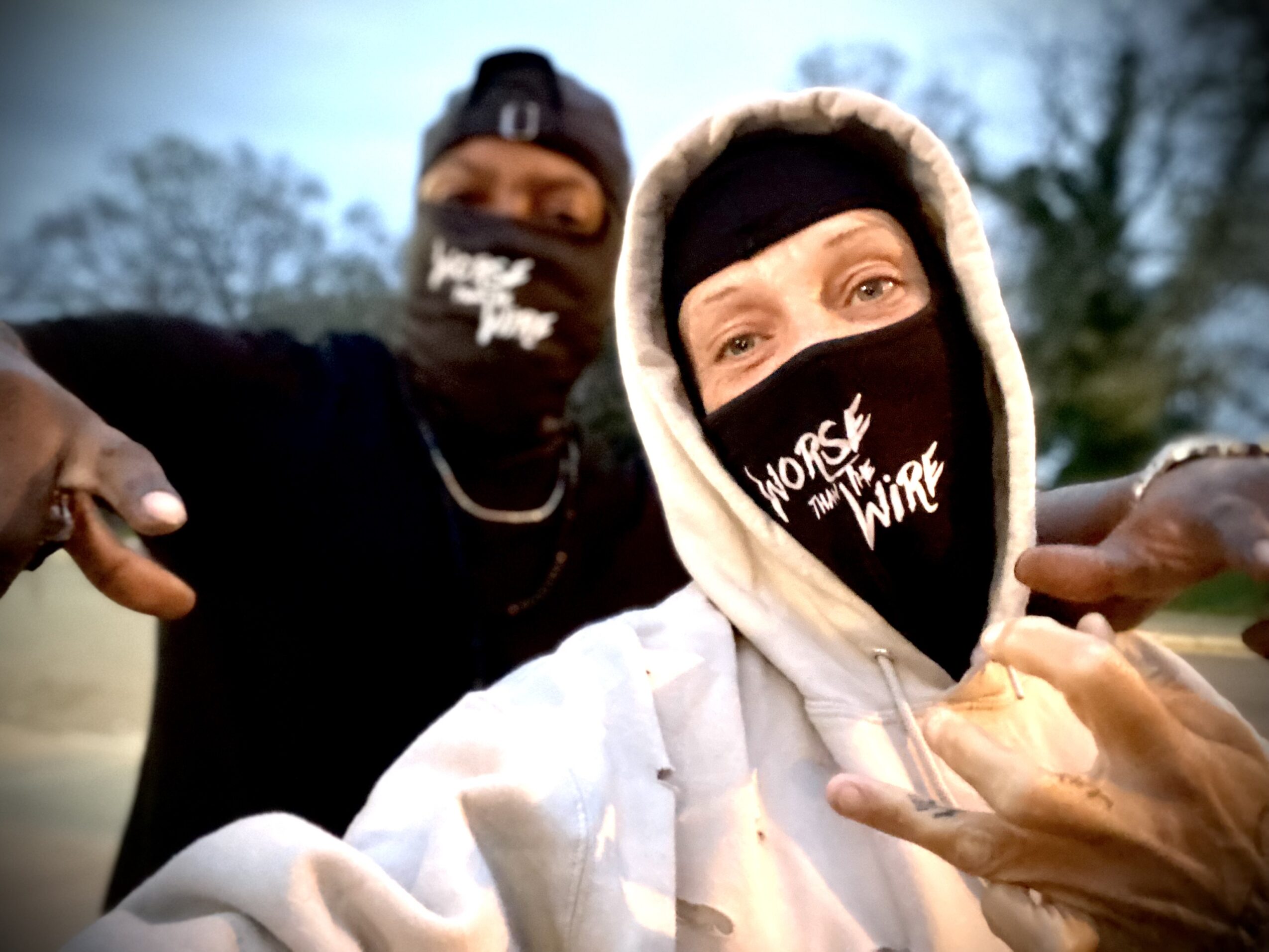 Two people wearing face masks outdoors, one in a white hoodie and the other in black, with trees in the background.