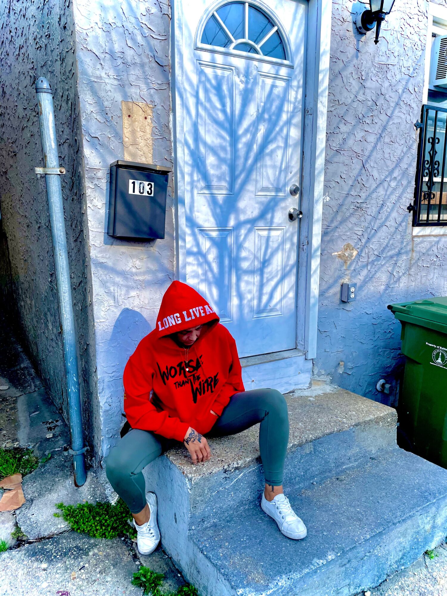 Person sitting on a concrete step in front of a white door, wearing a red hoodie, gray pants, and white shoes, with a shadow of a tree on the door.
