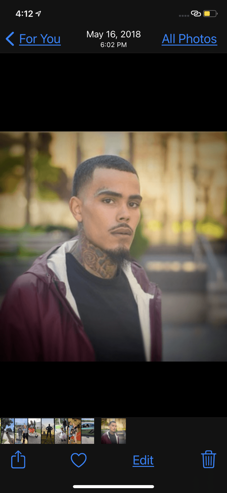 Young man with tattoos on neck and face, wearing a black shirt and maroon jacket, outdoors with blurred background.