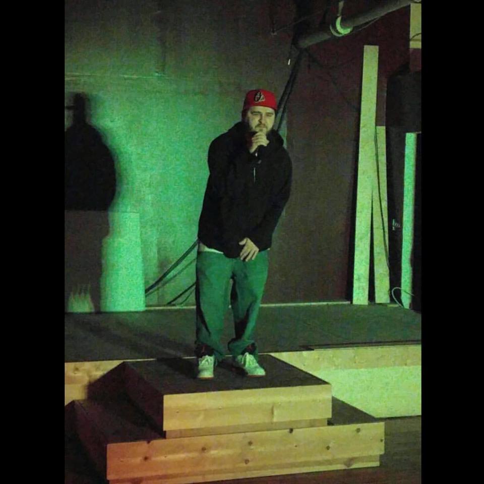 Person standing on a small stage holding a microphone, wearing a red cap and dark clothing, with a green-lit background.