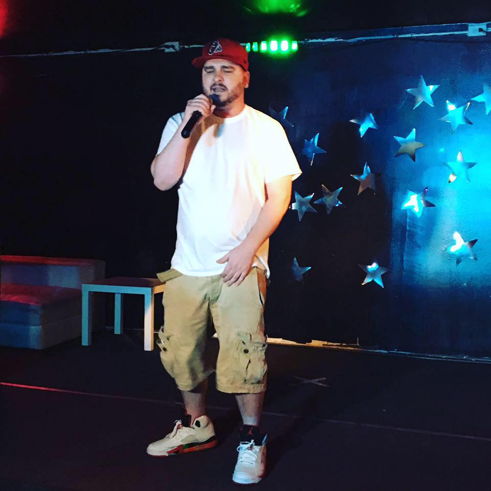 Man in white t-shirt, beige shorts, red cap, and sneakers holding a microphone on stage with star decorations and blue lighting