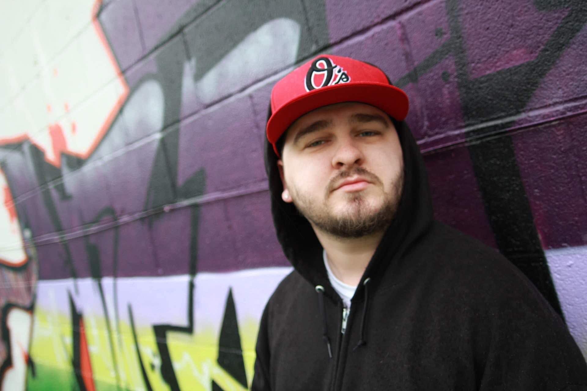 Young man with a beard and mustache wearing a red cap and black hoodie, standing in front of a colorful graffiti wall.