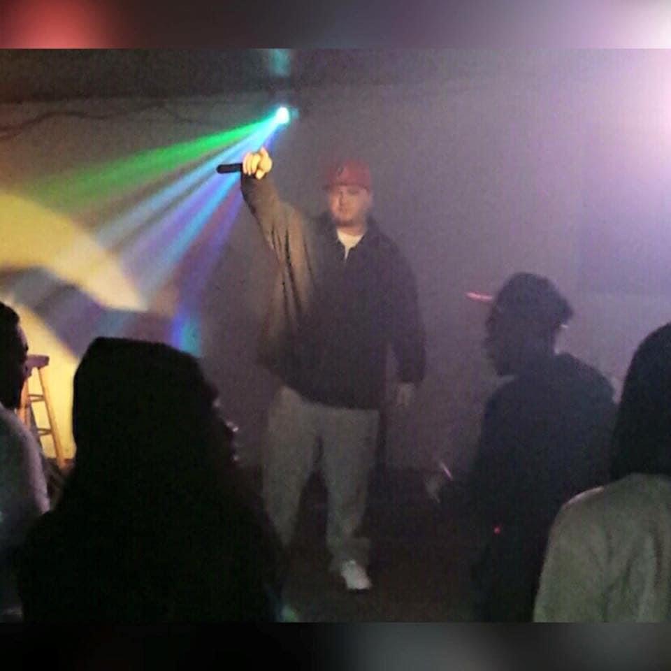 Person on stage raising fist, colorful lights, audience silhouettes, dark background.