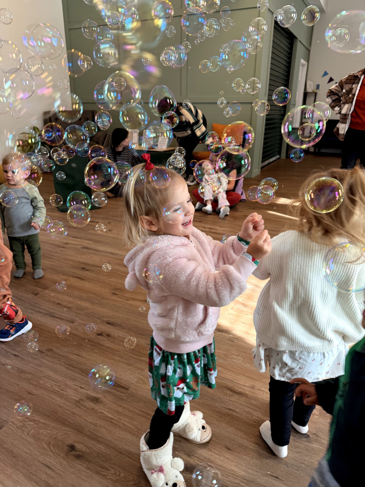 Children playing with bubbles indoors, some reaching for bubbles, others watching, with adults in the background, wooden floor, and decorated room.