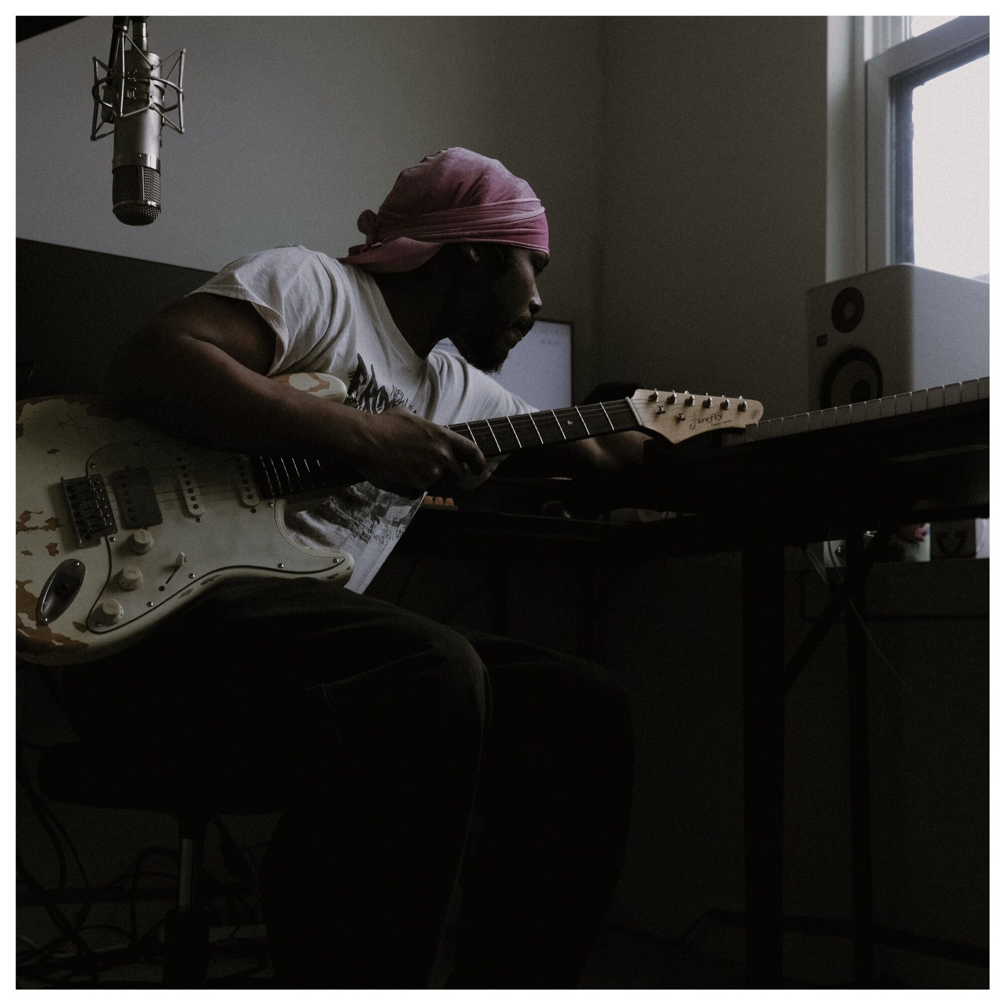 Person playing electric guitar in a dimly lit room with a window and audio equipment nearby.