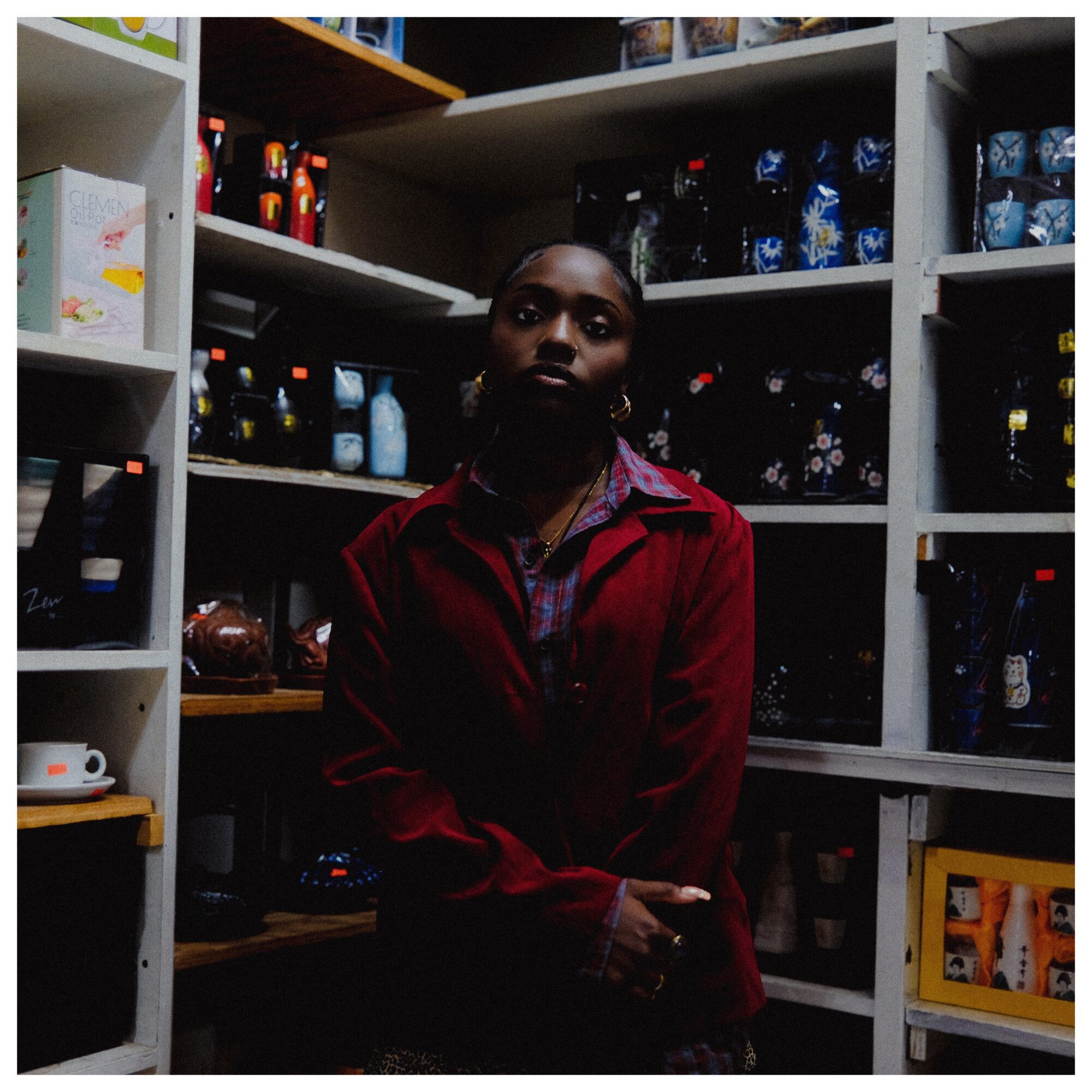 Person with dark skin and short hair in a red jacket stands in a store aisle with shelves of bottles and products behind them.