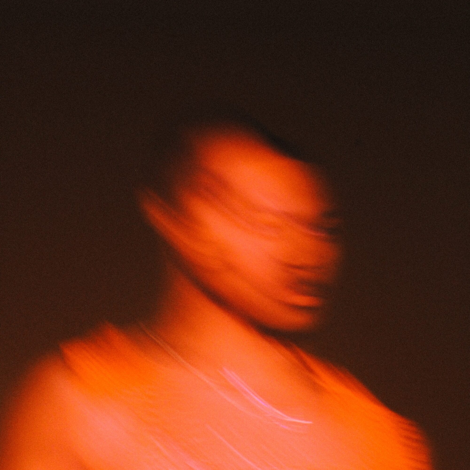Blurred image of a person with short hair, wearing an orange top, against a dark background.