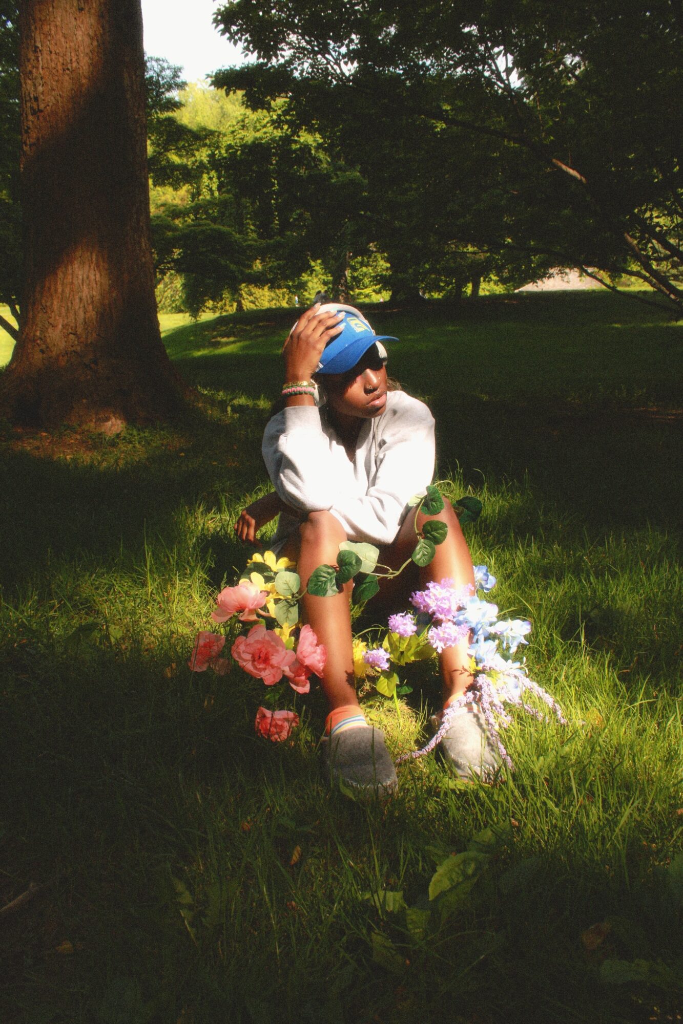 Child sitting on grass with flowers, wearing sunglasses and a blue hat, in a park with trees and sunlight.