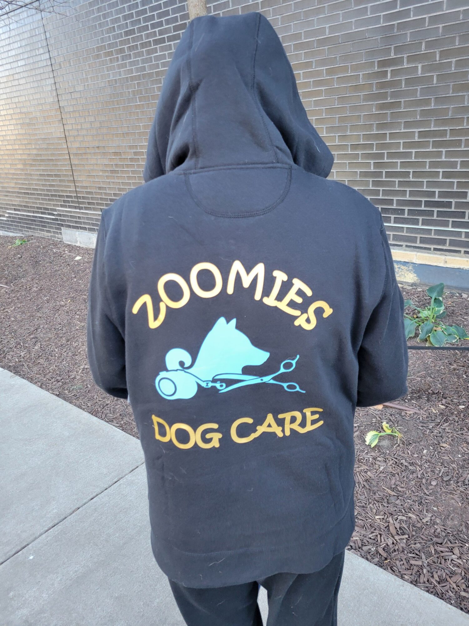 Person wearing a hoodie with a dog grooming logo, standing outdoors on a sidewalk.