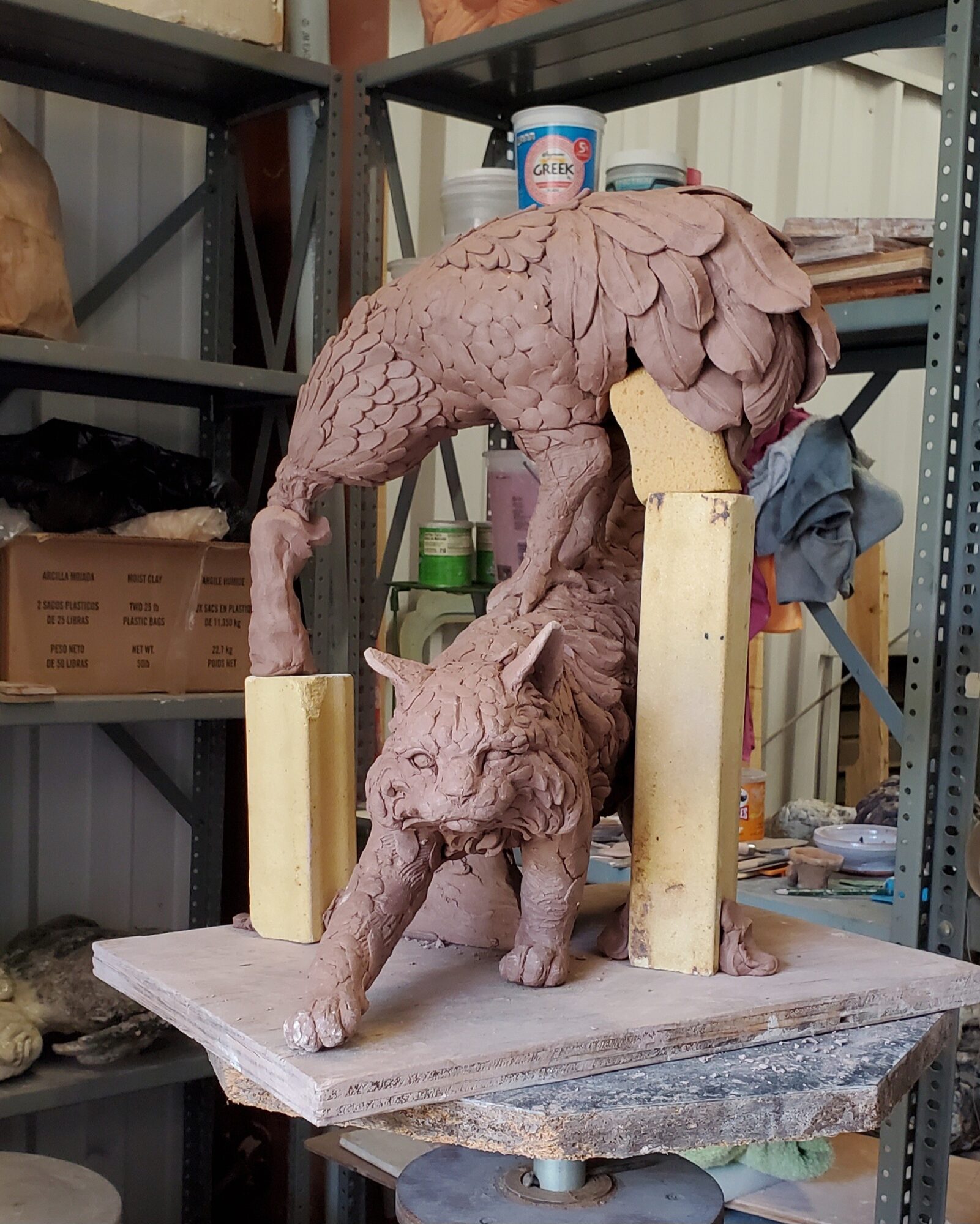 A sculpture of a creature with the body of a bird and the head of a cat, on a wooden base in a workshop.