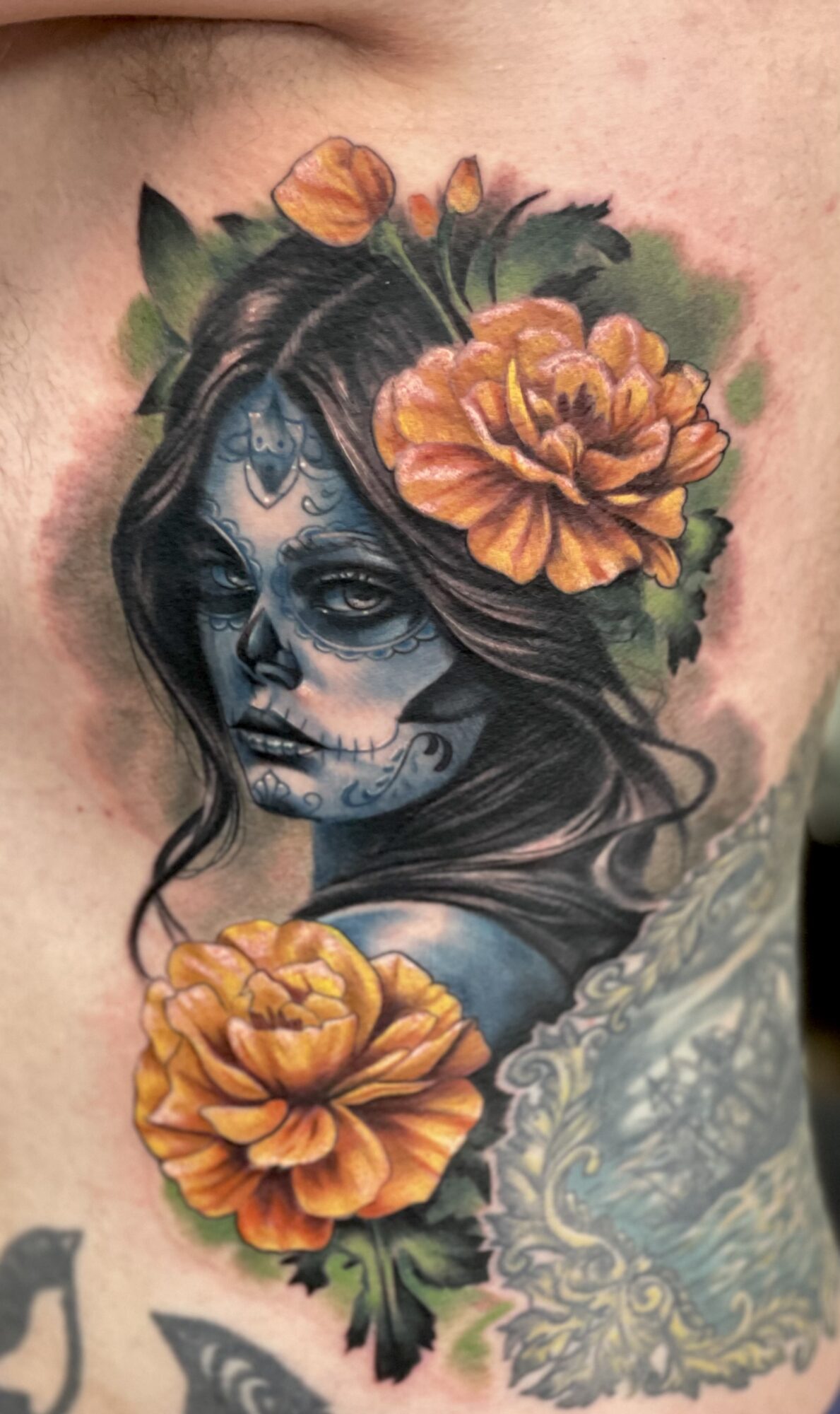 Tattoo of a woman with dark hair, decorated with orange flowers and intricate face designs, on skin.