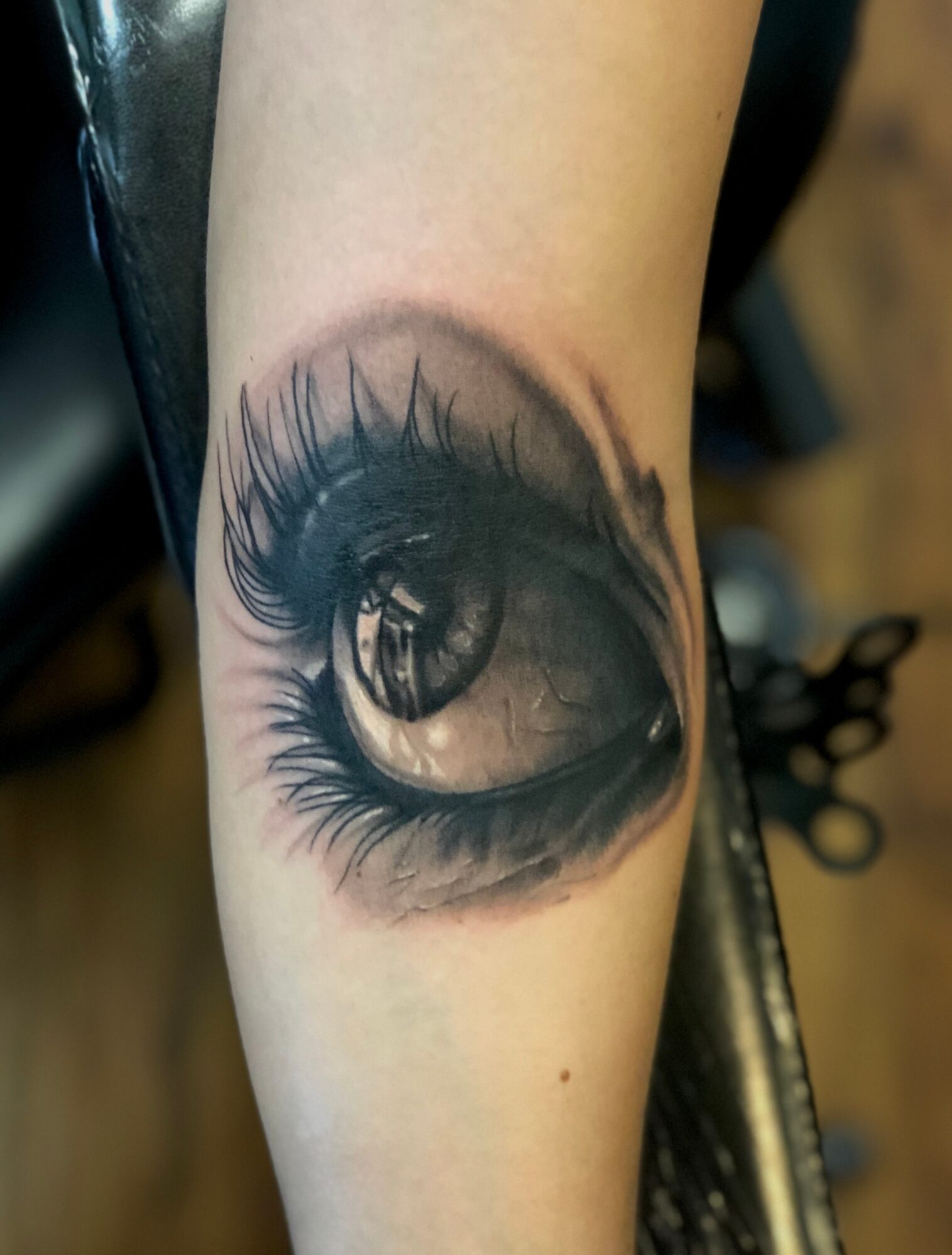 Realistic eye tattoo on person's arm with detailed eyelashes and iris, in black and gray ink.