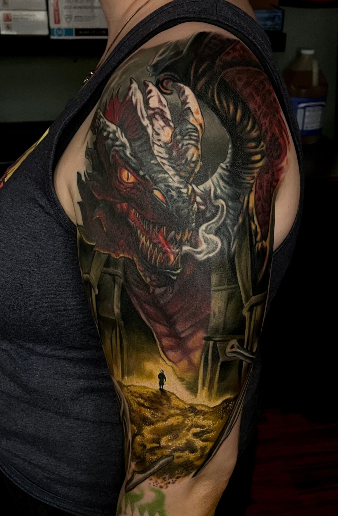 Tattoo of a dragon's head with smoke, above a landscape with a person and a dog, on person's upper arm.