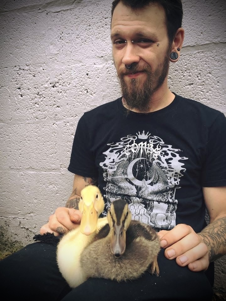 Man with beard and tattoos holding two ducks, one yellow and one gray, in front of a white wall.
