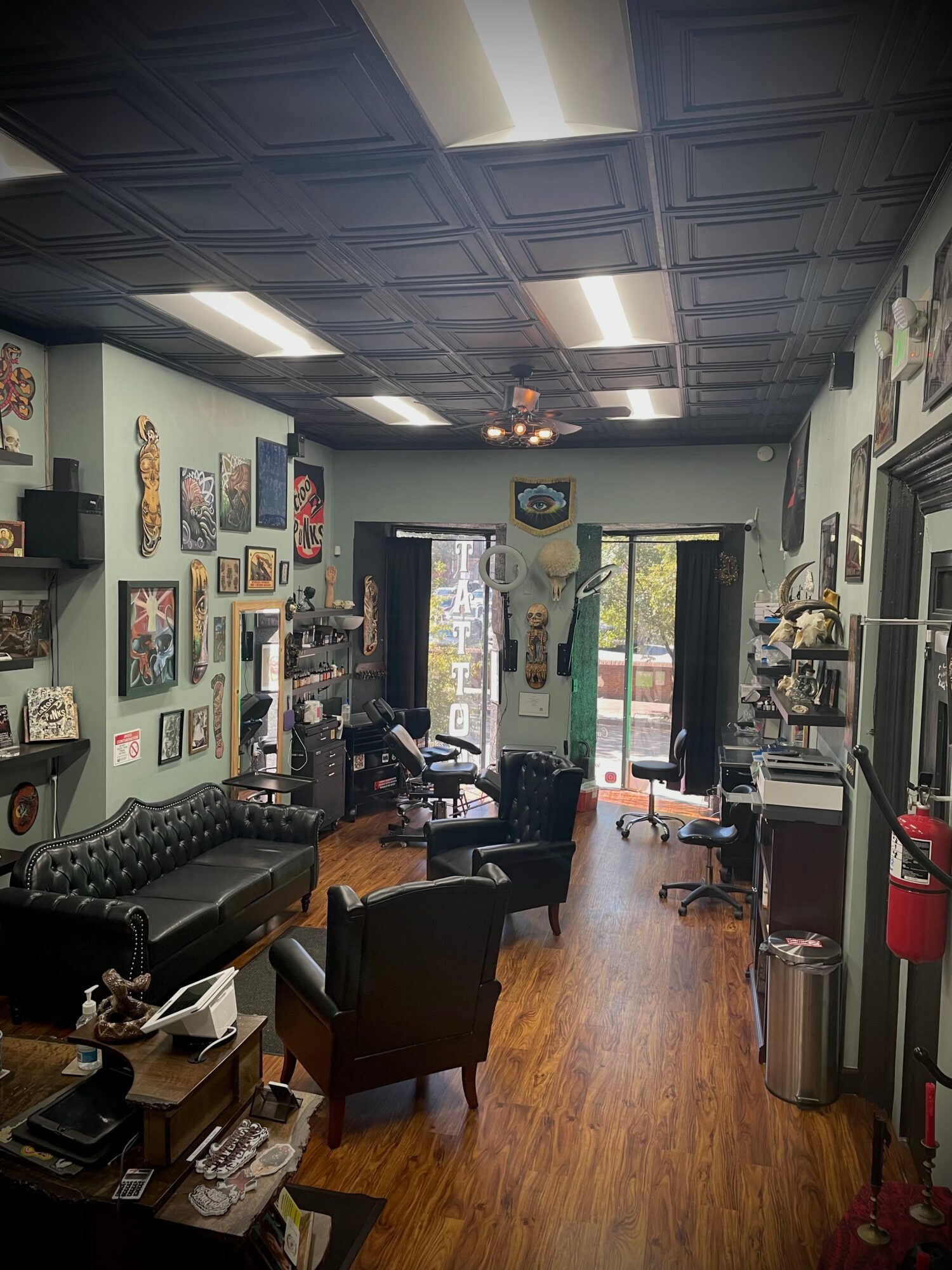 Room with wall art, seating, and large windows, ceiling with patterned tiles, hardwood floor, and various furniture and decor.