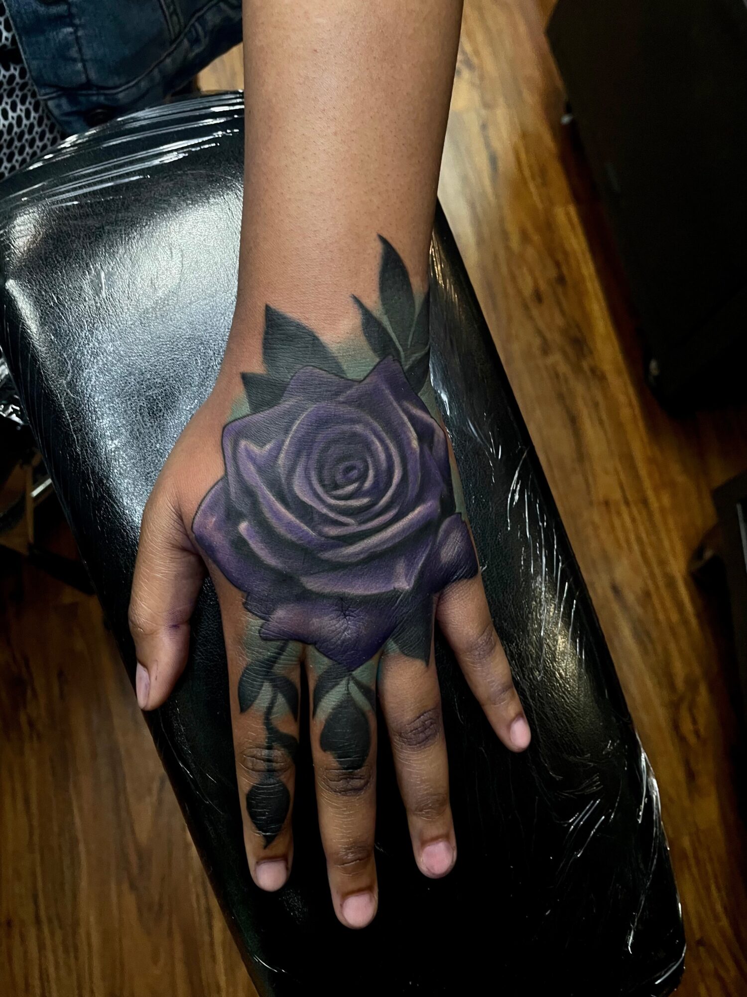Hand with a purple rose tattoo on the back, dark leaves, and a black background, resting on a black surface.