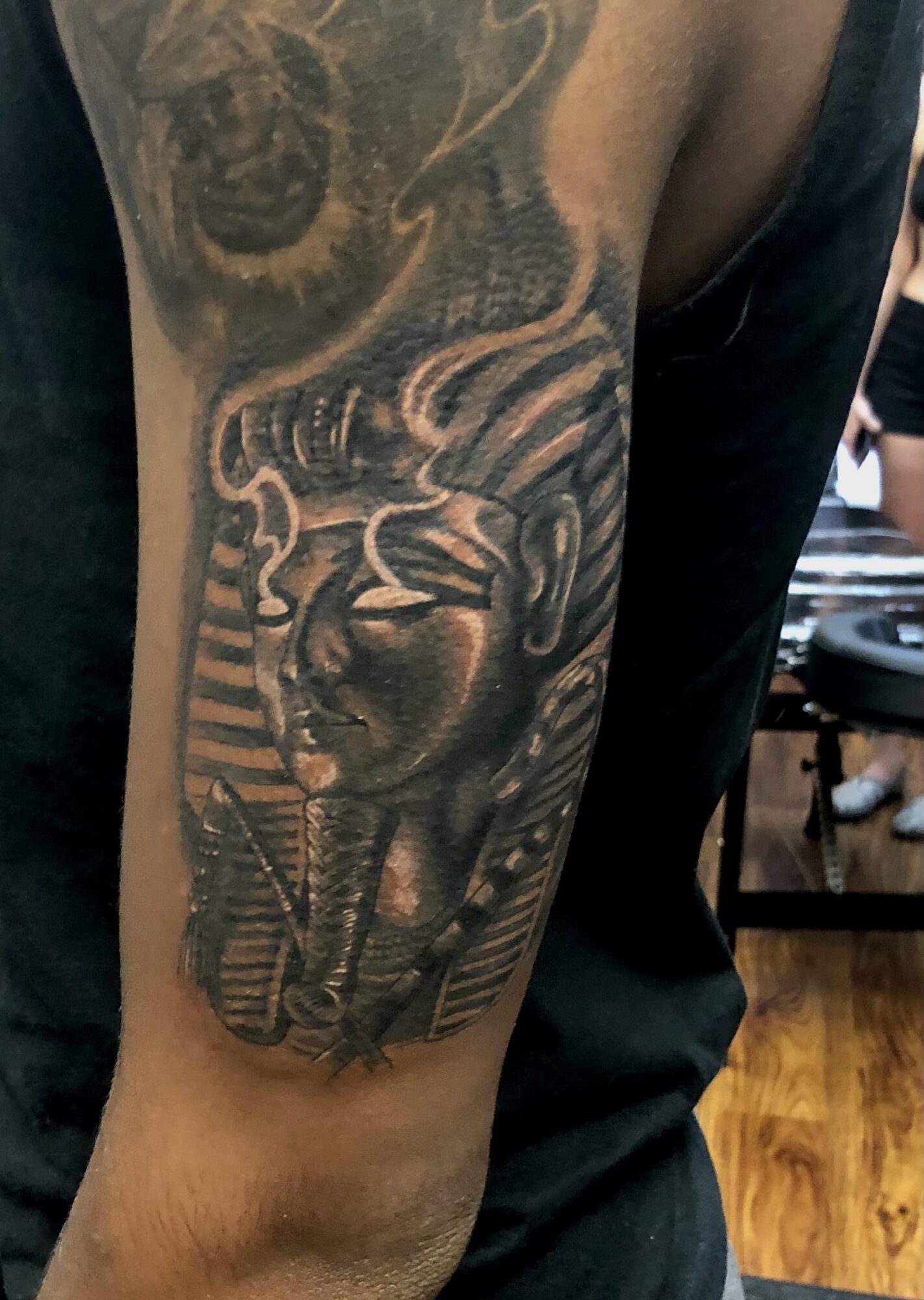 Black and gray tattoo of a woman's face with closed eyes, surrounded by abstract and striped patterns on upper arm.