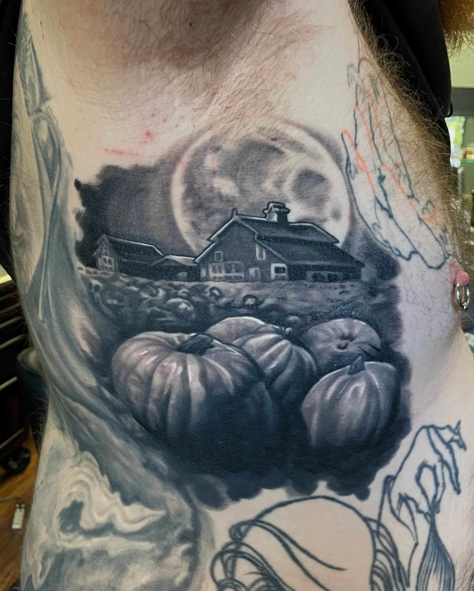 Black and gray tattoo of pumpkins, a house, and a moon on a person's chest, with some outlines and colors visible.