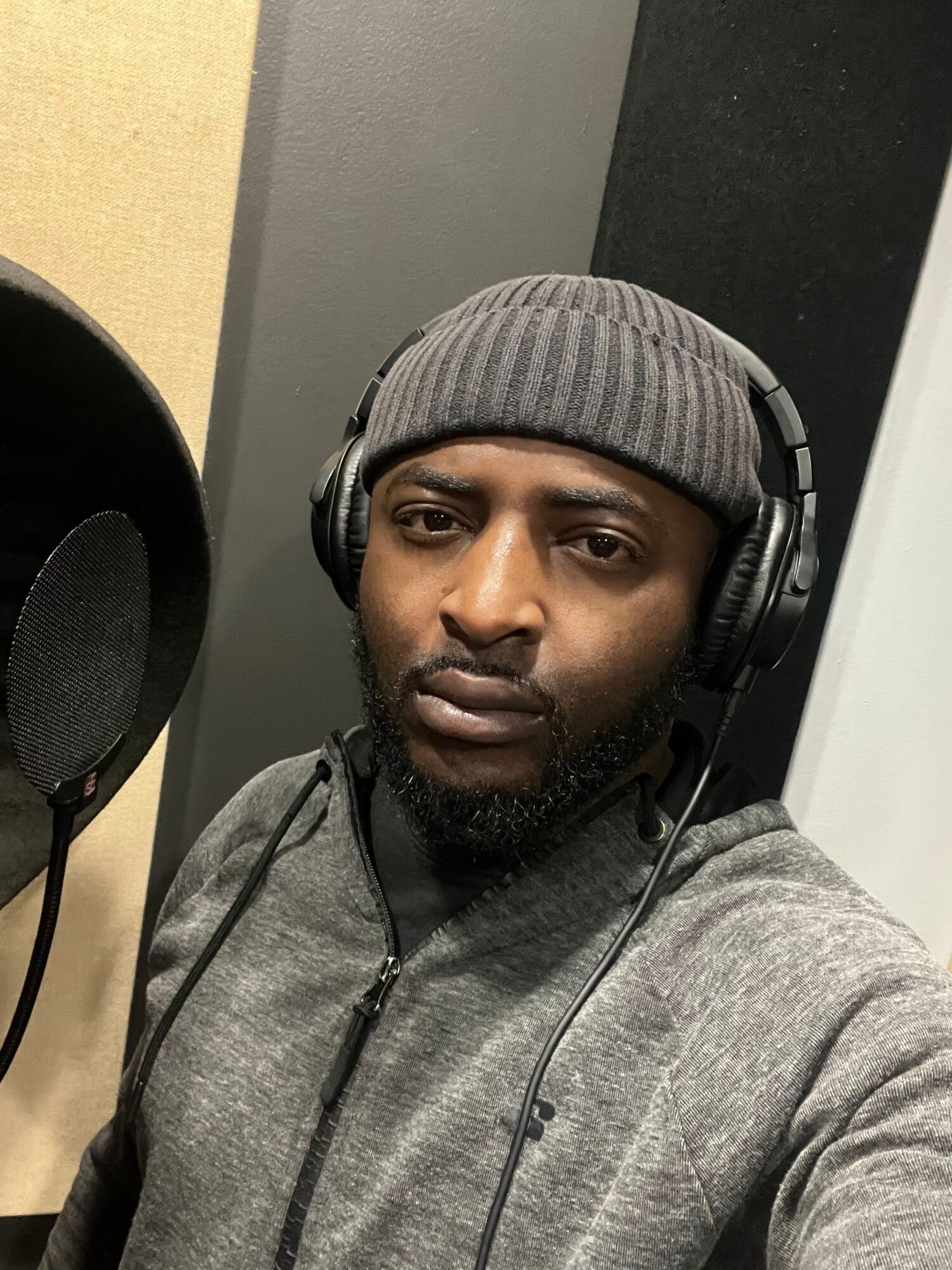 Man wearing headphones and a gray beanie, looking at the camera, in a room with black and beige walls.