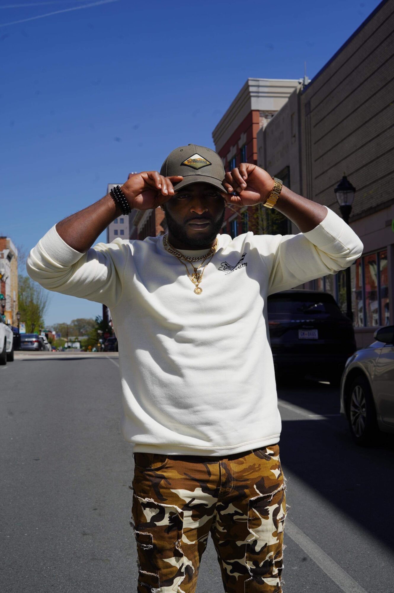 Man adjusting cap on street with buildings and cars, clear blue sky, wearing white sweatshirt and camouflage pants.