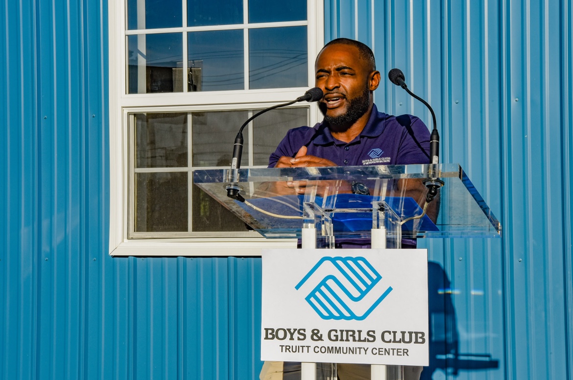 Man speaking at a podium with a Boys & Girls Club sign, blue building exterior, window, and blue curtains in background.