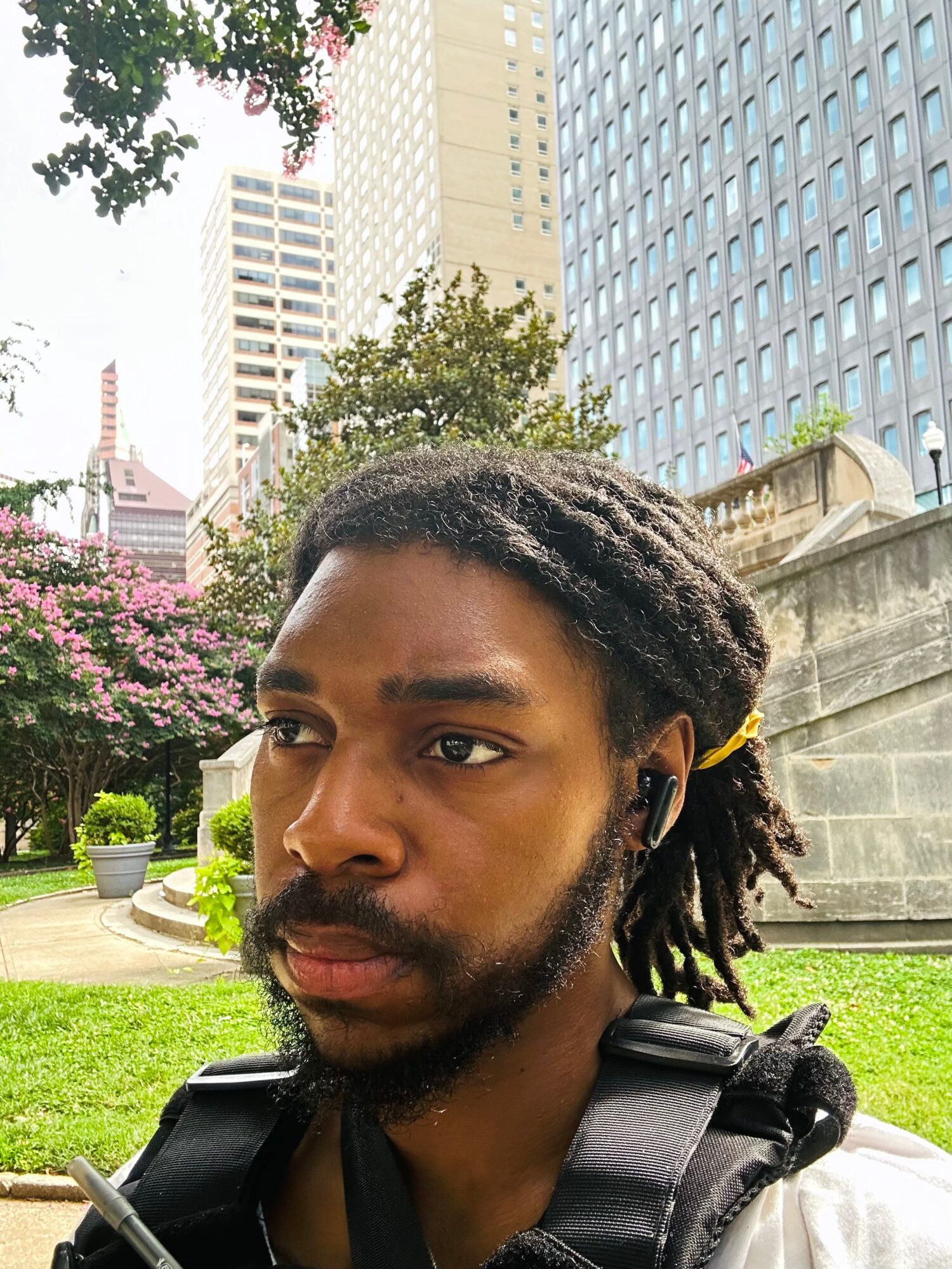 Young man with dreadlocks and a backpack outdoors in an urban park, buildings and trees in background.