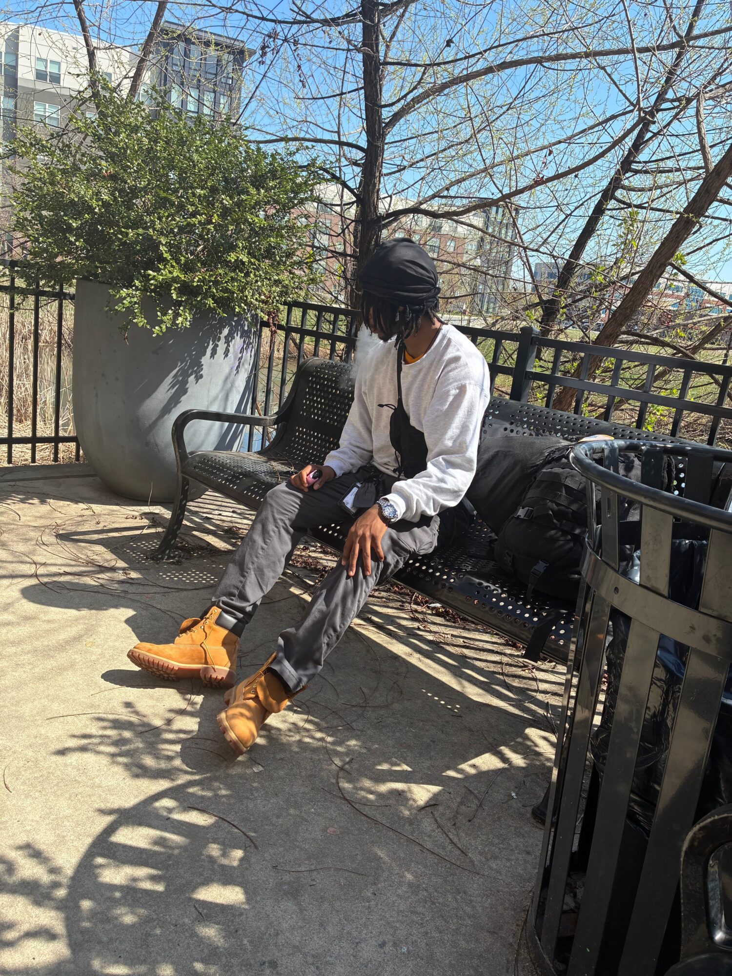 Person sitting on a bench outdoors, wearing a white jacket, gray pants, and tan boots, with trees and buildings in background.