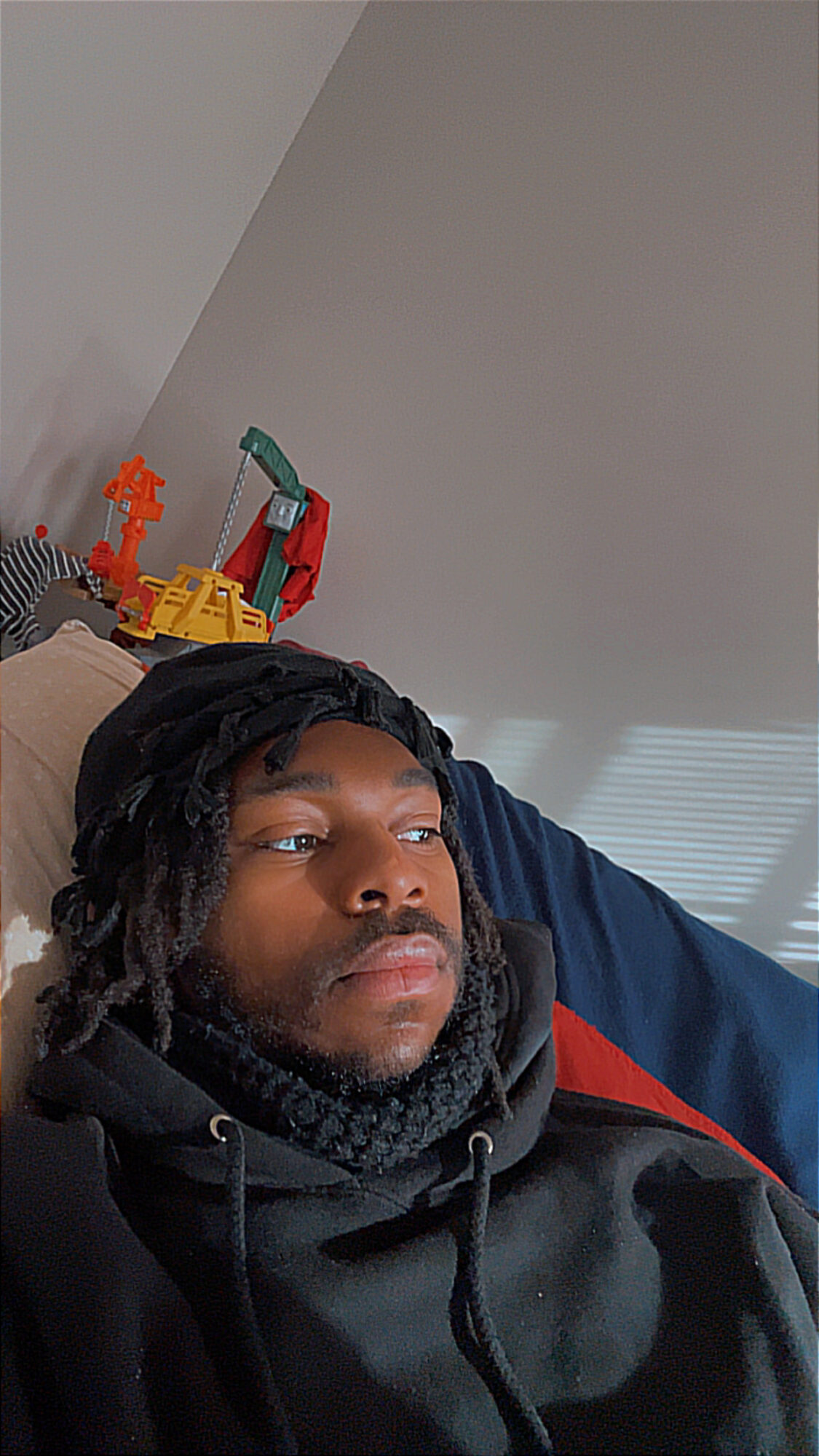 Person with dreadlocks lying on a bed, looking to the side, with toys on a shelf behind them.