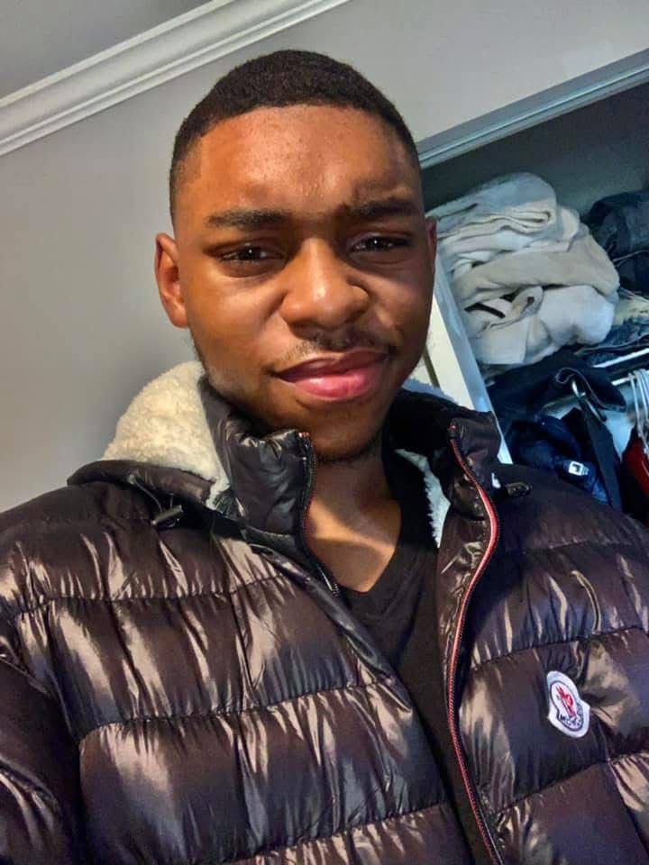 Young man with short hair wearing a black puffer jacket, standing indoors with a closet of clothes behind him.
