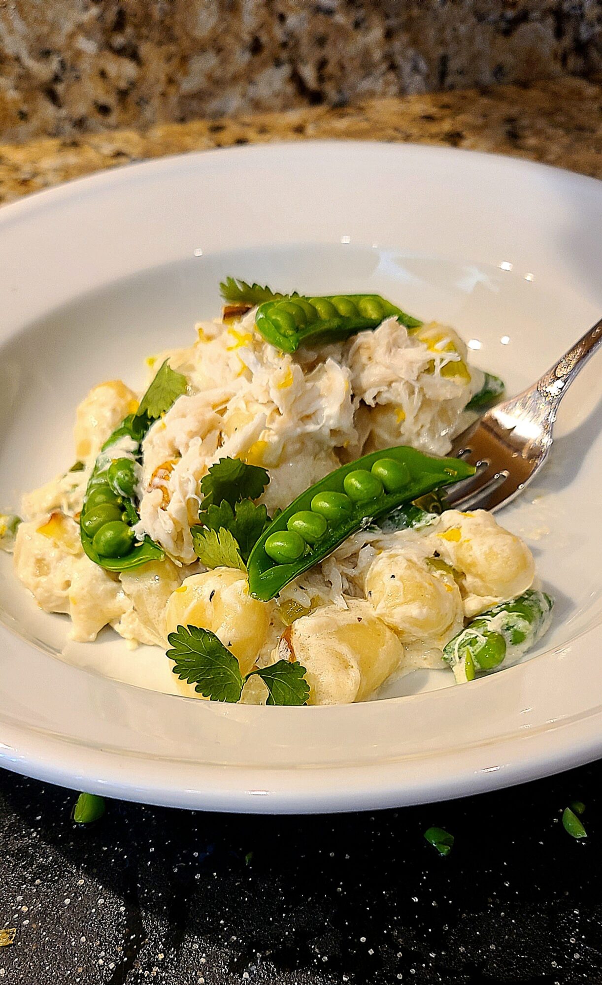 Creamy pasta with green peas and cilantro on a white plate, with a fork partially inserted, on a dark countertop.