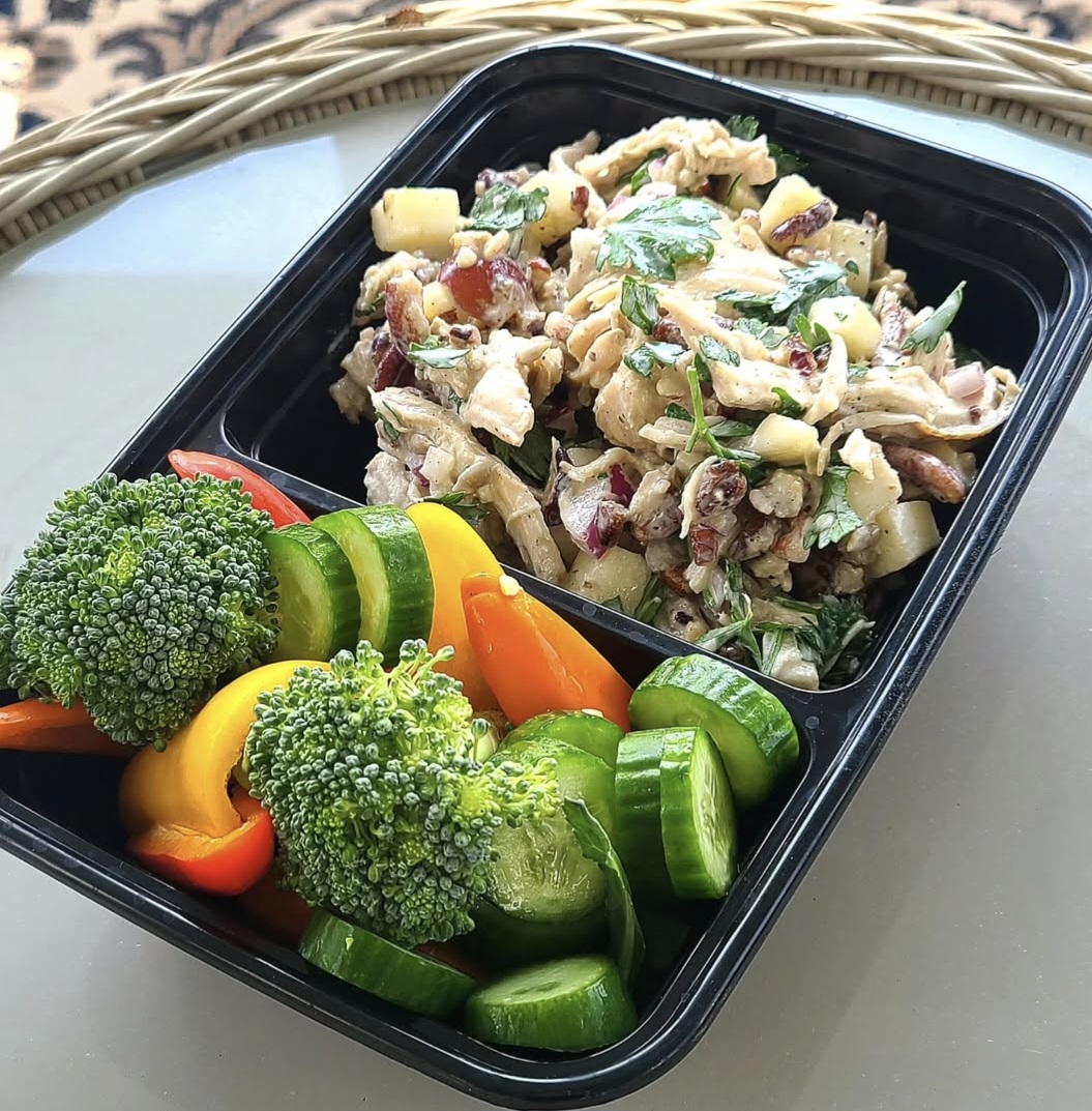 Black container with chicken salad and fresh vegetables including broccoli, cucumber, and bell peppers on a white surface.