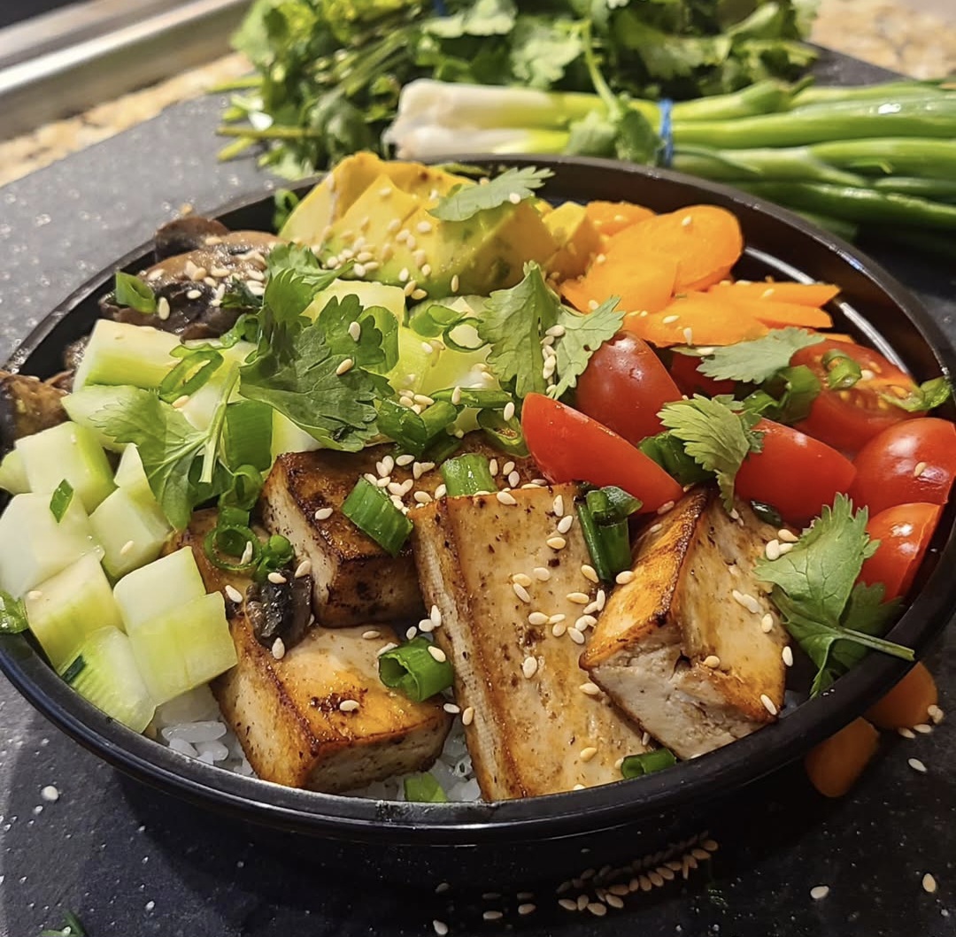 Bowl with grilled tofu, cherry tomatoes, cucumber, avocado, and herbs, with green onions and vegetables in the background.