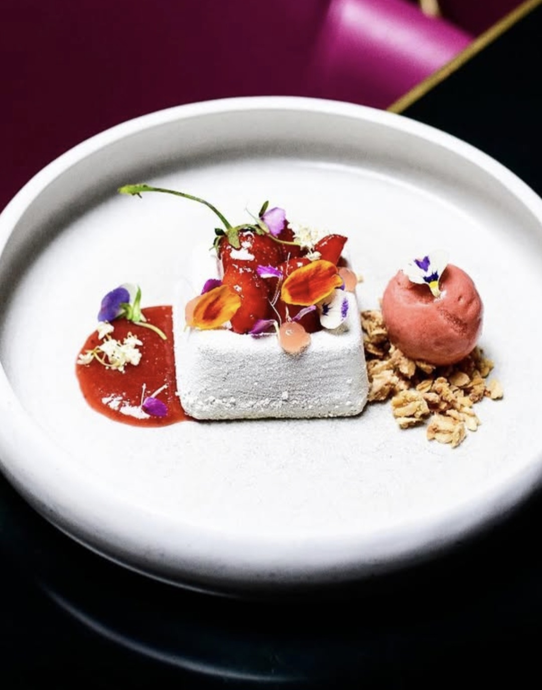 Elegant dessert plate with a white square cake, pink mousse, edible flowers, and granola on a white dish.
