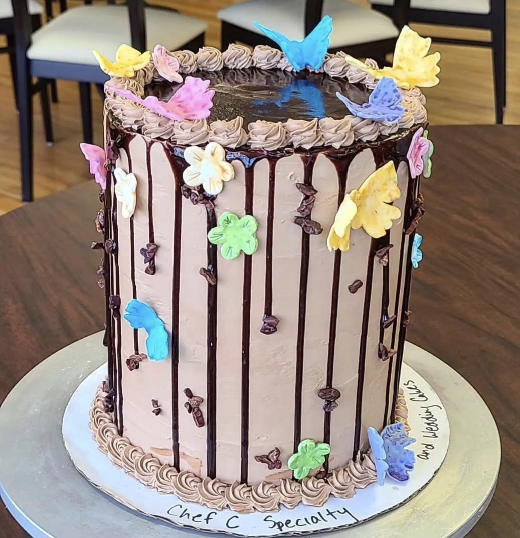 Tall chocolate cake decorated with colorful butterfly and flower decorations, topped with a blue butterfly, on a white base.