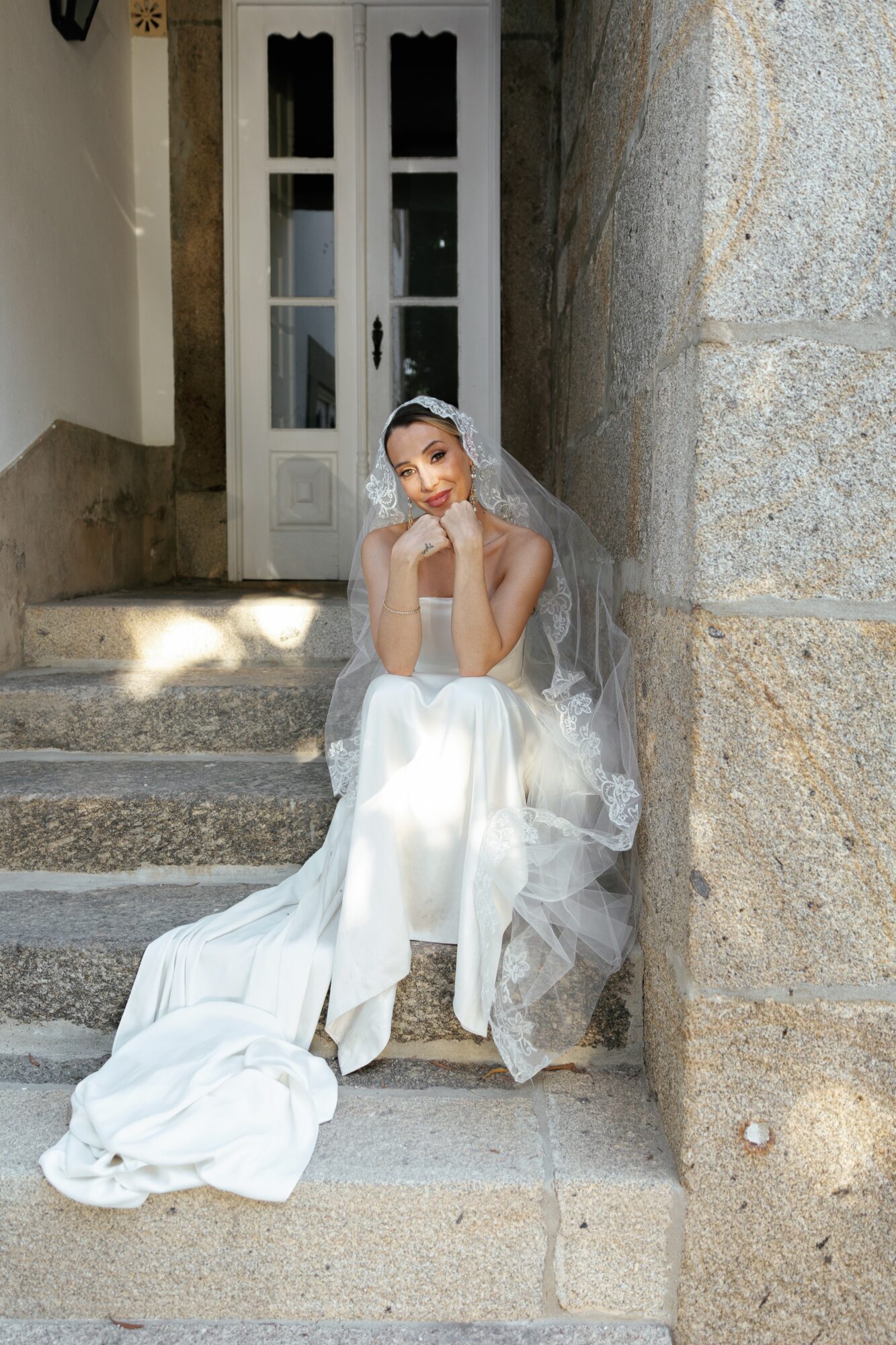 Young woman in wedding dress sitting on steps outside, leaning against stone wall, with a white door behind her.