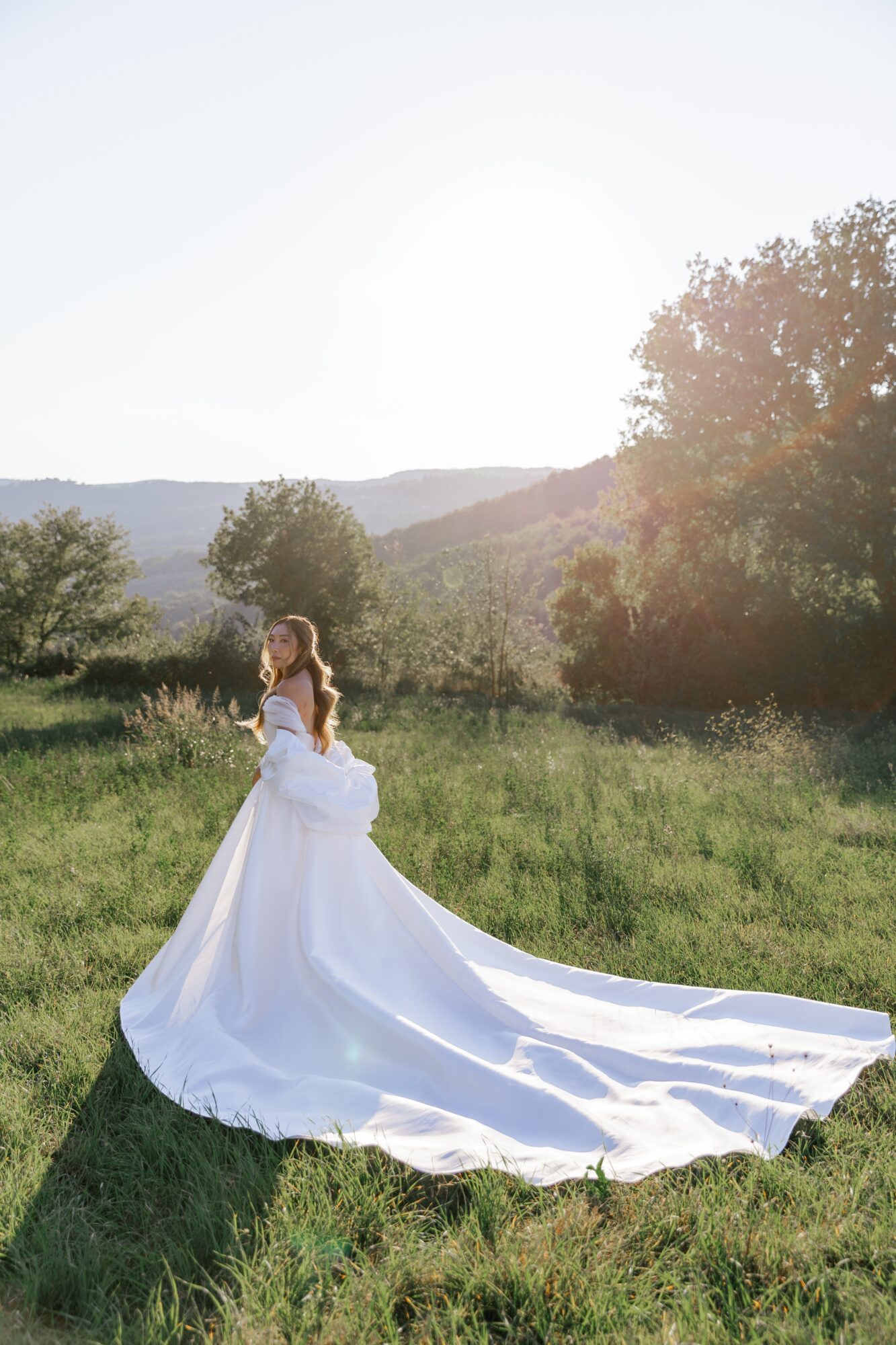 Woman in a white wedding dress standing in a grassy field with trees and hills, sunlight shining brightly behind her.