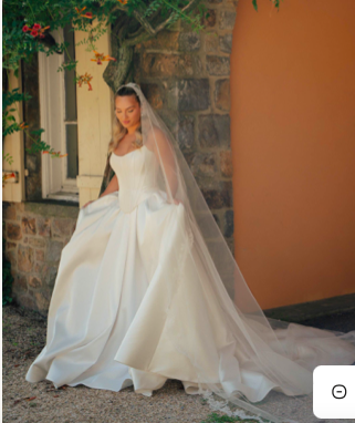 Bride in white wedding dress with veil, standing outdoors near a stone wall and window, holding her dress.